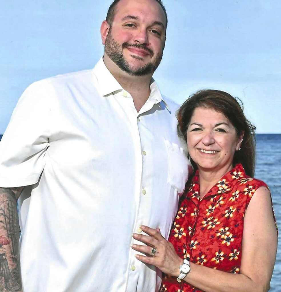 A man in a white shirt and a woman in a red floral sleeveless top stand close together, smiling, with the ocean and blue sky in the background.