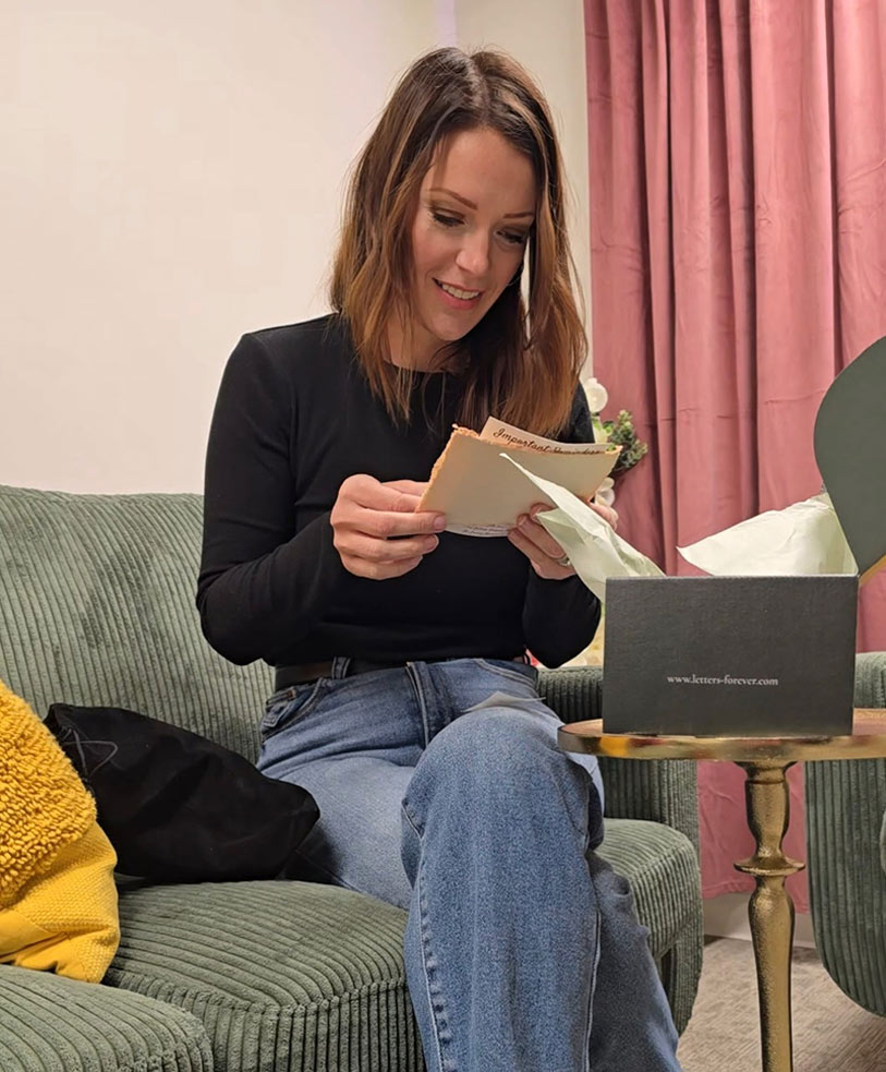 A woman with brown hair in a black top sits on a green couch, smiling as she reads letters from the Memory Vault, a gray box on a small table. A yellow pillow and pink curtains are seen in the background.