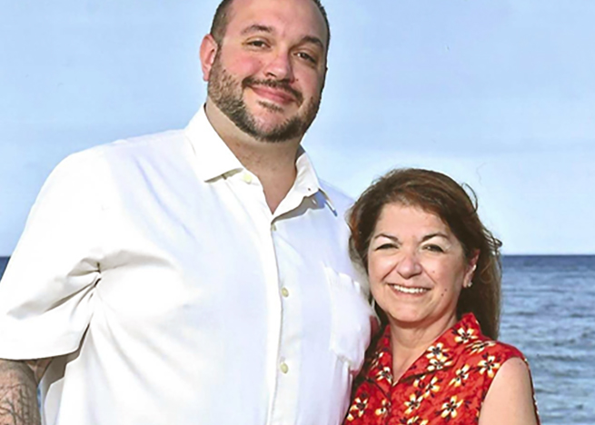 A man and woman stand smiling together in front of the ocean under a clear blue sky. The man wears a white shirt, and the woman wears a red patterned sleeveless blouse.