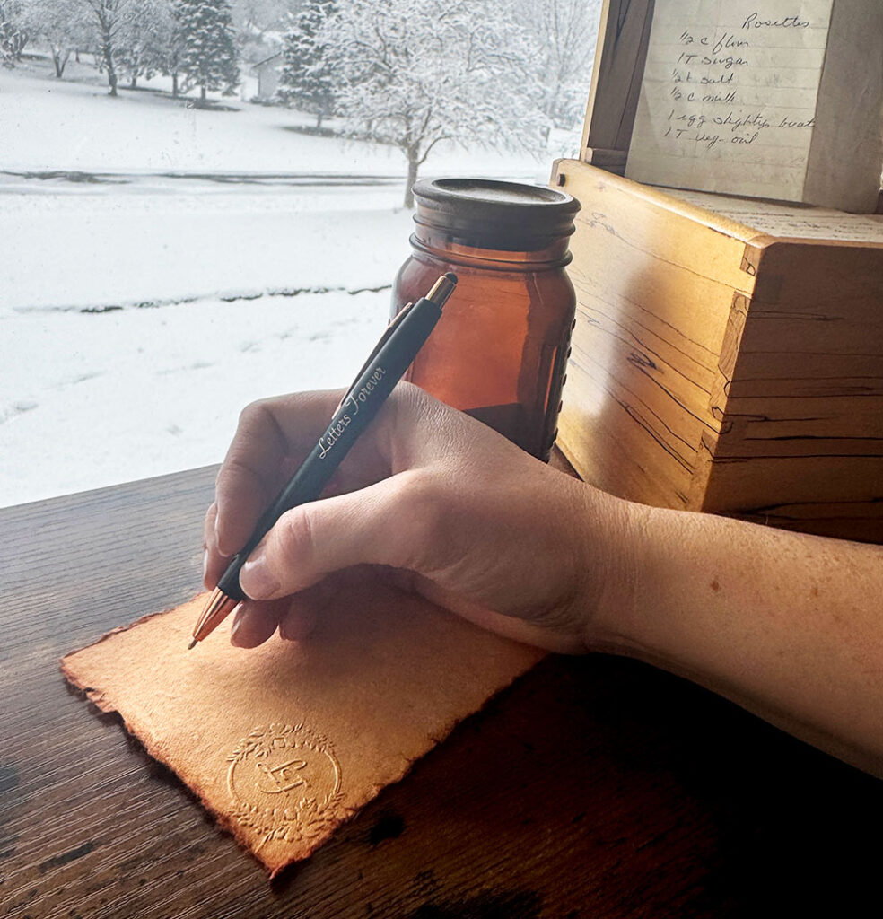 A hand holding a pen writes on a piece of parchment paper at a wooden desk. A brown jar, stacked books, and snowy trees outside a window are visible in the background.